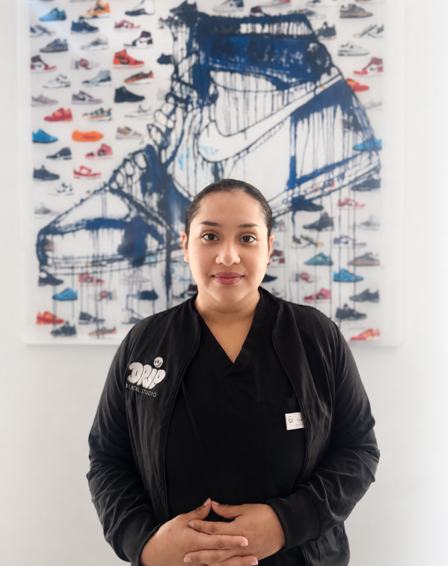 Portrait of Francis Registered Dental Assistant with dark hair pulled back, wearing a black Drip Dental Studio jacket and standing in front of sneaker artwork.