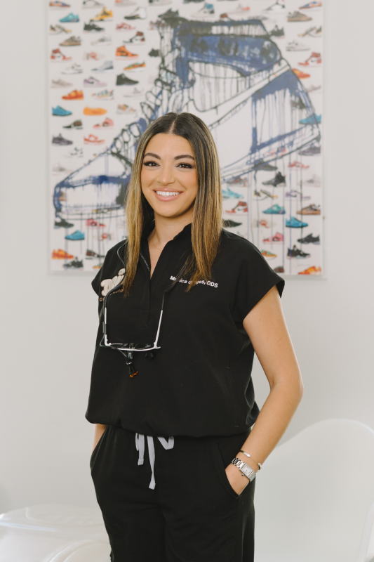 Portrait of Dr. Monica Gerges with dark hair in black scrubs with dental loupes, posing in front of sneaker wall art.